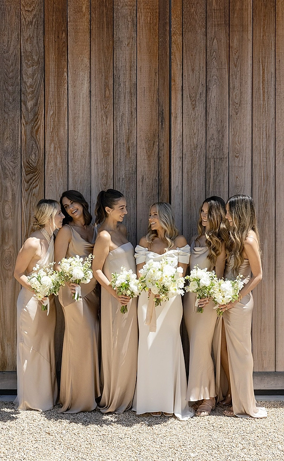 Bridesmaid group photo with bridesmaids holding bouquets, neutral dresses and ribbon wraps posed by a rustic wood wall on gravel ground