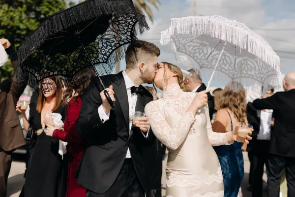 Wedding kiss as the newlyweds kissing under black and white fringed lace parasols, holding champagne coupes amid outdoor guests and palms