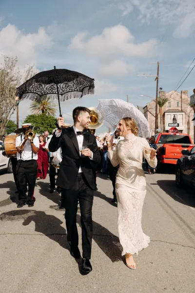 Wedding second line with bride in lace dress and groom in tuxedo under a black fringe parasol, marching behind a brass band on a city street