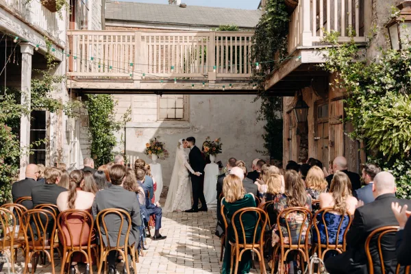 Ceremony kiss under a flowered wedding arch with string lights, bride in lace dress and veil, groom in black suit in ivy courtyard