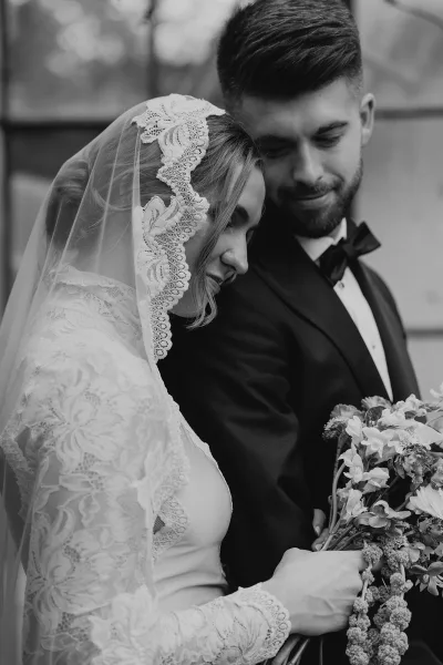 Couple portrait in black and white, bride leaning on groom in tuxedo, lace veil and sleeves, holding a bouquet by foliage and building exterior