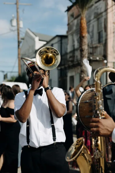 Wedding band leading a brass band wedding parade, musicians in bow ties and suspenders playing trombone and sousaphone on a sunny street with crowd and palms
