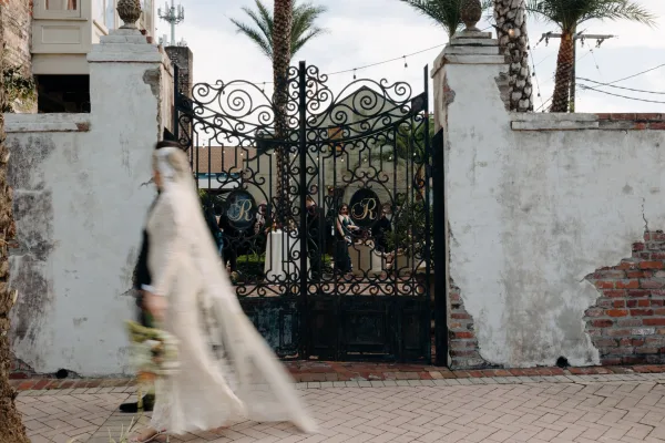 Bride walking with bouquet, wedding dress and veil trailing past a wrought iron gate under string lights in a palm-lined courtyard