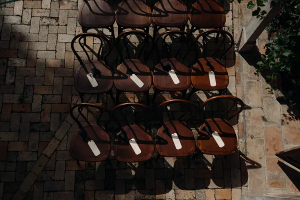 Ceremony seating with wedding chair tags on wood bistro chairs, black tassels draped over backs on a sunlit brick patio courtyard