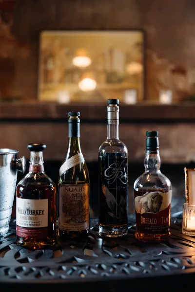 Wedding whiskey bar with bourbon bottles, rocks glasses, and an ice bucket, lit by votive candles beside a framed sign in an indoor lounge