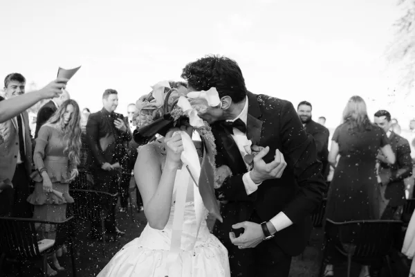 Confetti exit as newlyweds share a ceremony exit kiss, bride clutching a bouquet while guests line the aisle in celebration.