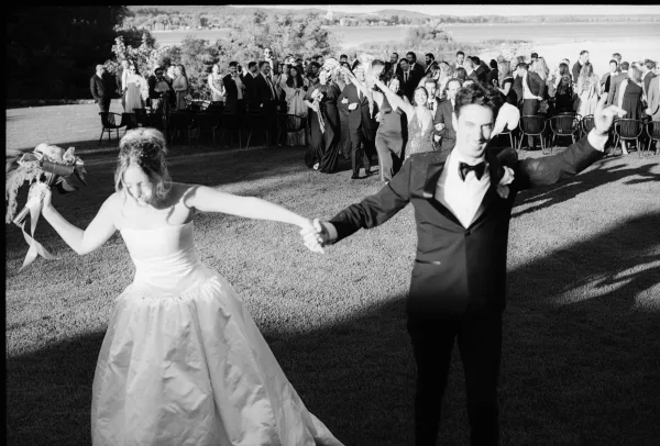 Wedding recessional with a couple holding hands, the bride lifting a ribboned bouquet, walking past cheering guests on a grassy lawn by the water.