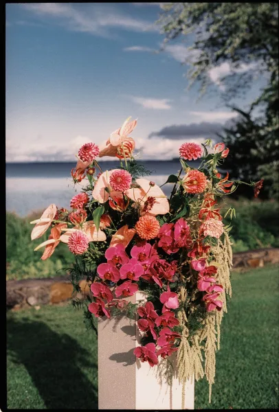 Ceremony flowers in a pedestal wedding arrangement featuring lush orchids, set on a lakeside lawn with trees and a stone wall beyond