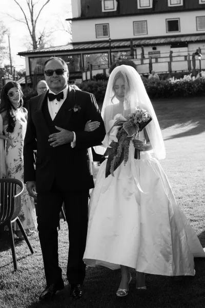 Ceremony processional with father escorting bride down an outdoor lawn aisle, her bridal veil trailing as guests watch by trees and chairs.
