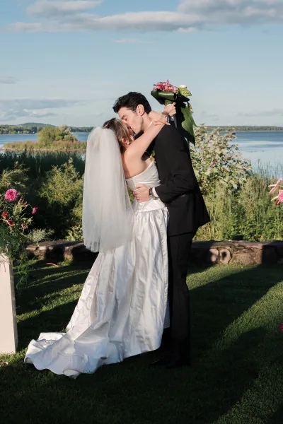 Wedding kiss portrait capturing the first kiss moment as the bride and groom embrace by a lakeside lawn, her pink bridal bouquet lifted.