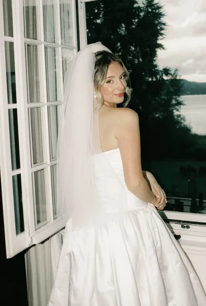 Bridal portrait of a bride by window, glancing over her shoulder in a strapless satin gown, framed by open curtains with lake and trees beyond.