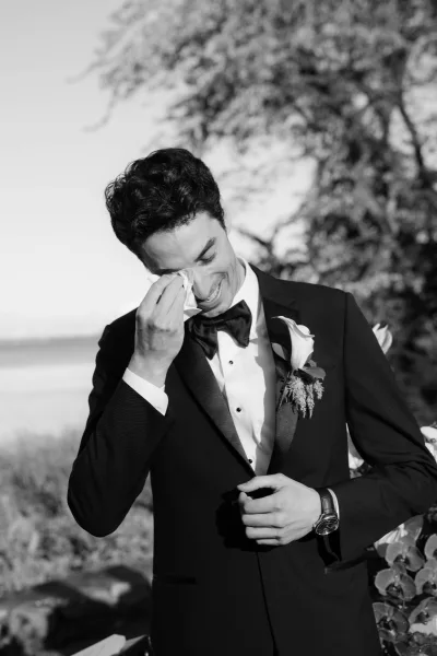 Black and white groom portrait capturing an emotional groom reaction as he dabs a handkerchief, set against trees and water.