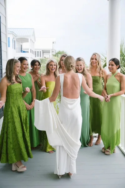Bridesmaid reveal as the bride in a low back gown shows her look to bridesmaids in green dresses on a porch with white columns