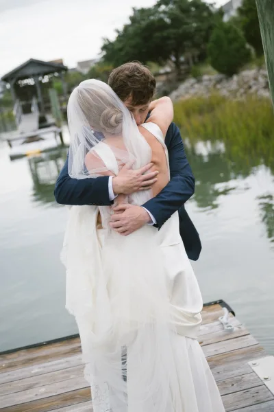 Wedding couple portrait of a bride and groom hug on a wooden dock by calm water, her veil trailing over a satin gown under overcast sky