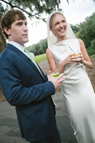 Wedding cocktail hour with bride holding cocktail and groom holding drink, laughing on a lawn path beneath trees and open sky