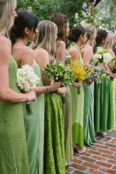 Bridesmaids lineup in green bridesmaid dresses holding white, yellow, and purple bouquets with greenery on a brick walkway in a garden