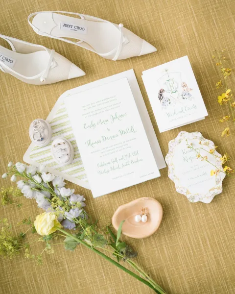 Invitation suite wedding invitation flatlay with envelopes, vow book, rings, pearl drop earrings, white heels and yellow-blue flowers on tan linen