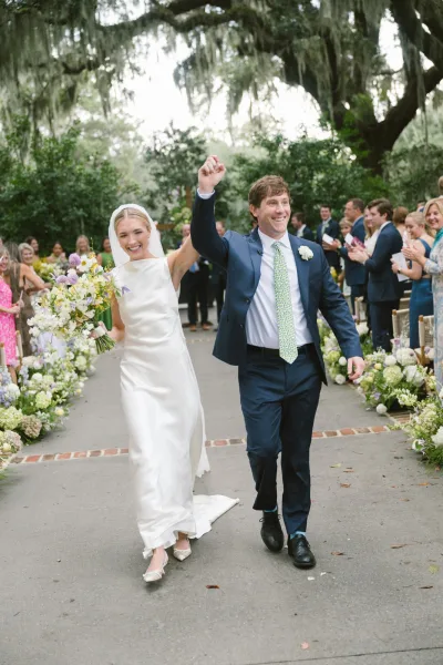 Wedding recessional just married walk as bride holds bouquet and groom in navy suit exit past cheering guests under mossy trees on walkway