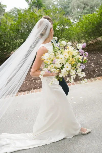 Bride and groom portrait with bride with long veil flowing as they walk a garden walkway, her bouquet in hand and groom in navy suit