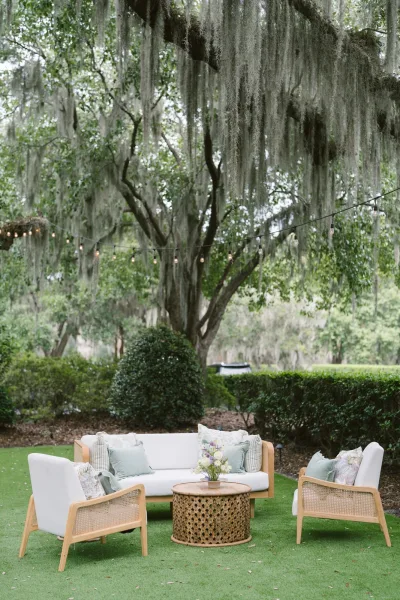 Outdoor lounge setup for a wedding lounge area with white sofa, cane chairs and floral centerpiece under string lights on an oak-shaded lawn