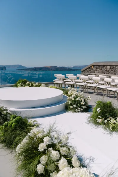 Ceremony setup featuring modern white ceremony decor with a round platform overlooking an ocean view, framed by glass railing and blue sky.