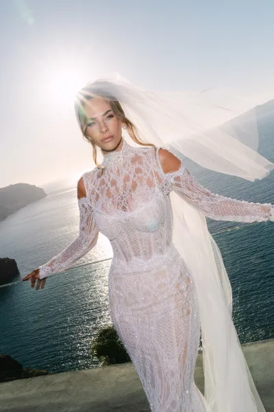 Bridal portrait of a bride in lace gown on a sunlit balcony above ocean cliffs, her long veil drifting as the sky frames the scene.