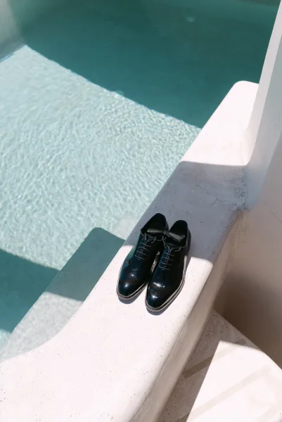 Groom shoes, sleek tuxedo shoes set beside a folded bow tie on a stone ledge, with shimmering pool water reflecting light behind.