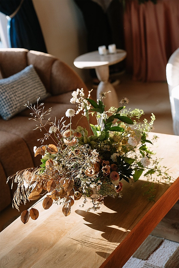 Wedding bouquet with wildflower wedding bouquet textures, greenery and dried grasses resting on a wood coffee table in sunlit room