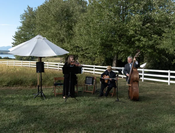 Wedding musicians performing as an outdoor wedding band with violin and double bass beside microphones on a grassy field by a white fence