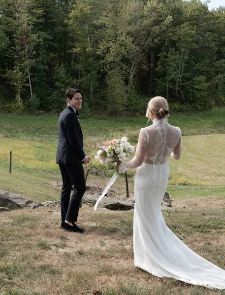 First look moment as bride in lace-sleeve gown approaches groom in black tuxedo, holding bouquet with ribbon in a meadow by forest trees