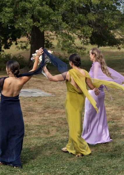 Bridesmaid moment as bridesmaids walk away in colorful dresses, holding bouquets with a chiffon scarf accent across a grassy field by a large tree