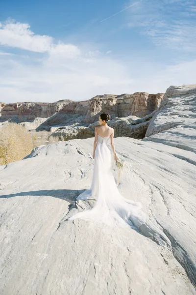 Bridal portrait of a bride in a back view wedding dress holding a bouquet with ribbon, walking toward desert cliffs under blue sky