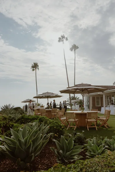 Cocktail hour setup with outdoor cocktail seating under patio umbrellas, arranged on a lawn with an ocean view and palm trees.