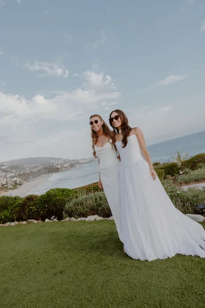 Couple portrait of two brides in sunglasses, smiling arm in arm on a coastal lawn with ocean and beach beyond under a clear sky.