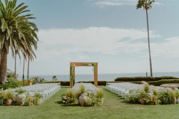 Ceremony setup for an outdoor wedding ceremony facing the ocean, with a wooden arch on a lawn bordered by palm trees.