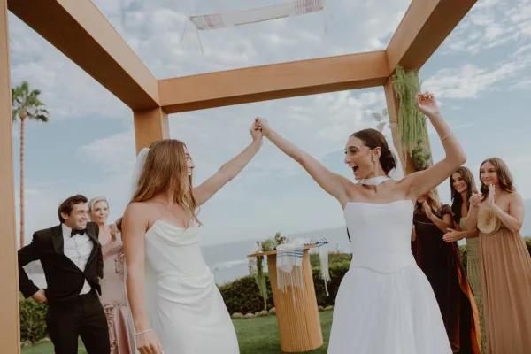 Ceremony moment with brides holding hands beneath a chuppah, joyful and newly married against an ocean backdrop with palm trees.