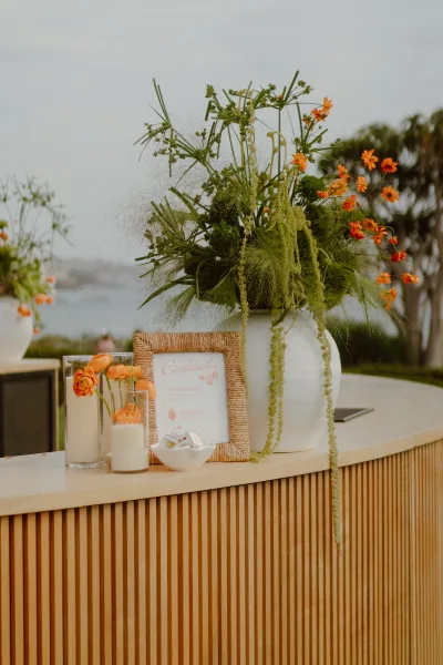 Wedding bar decor featuring a signature cocktail sign in a rattan frame, set against water, trees, and open sky.