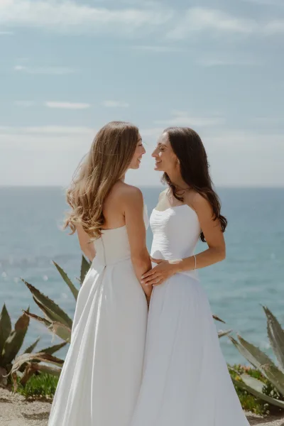 Couple portrait of a bride and bride holding hands by the ocean, one with a delicate bracelet, framed by open sky and coastal plants.