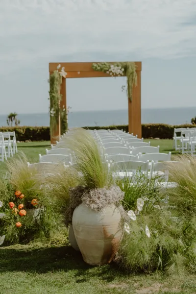 Ceremony setup for an outdoor ceremony aisle centered on a wooden arch, with the ocean and sky beyond a manicured lawn and hedges.