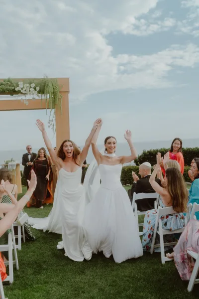 Ceremony recessional during their just married exit, two brides hold hands walking the aisle as guests cheer, ocean backdrop and wooden arch.