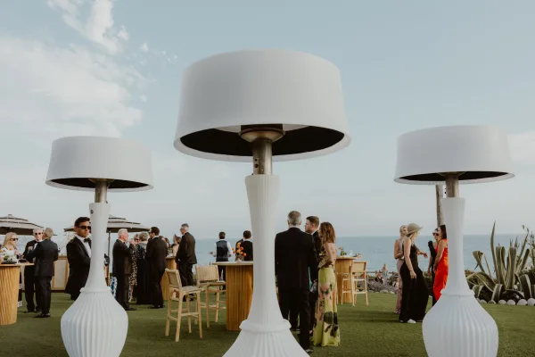 Cocktail hour setup for an outdoor cocktail reception with patio heaters on a lawn overlooking the ocean and a single palm tree silhouette