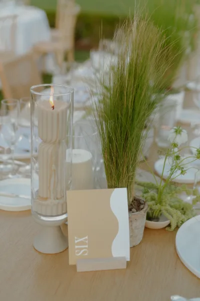 Wedding table number anchoring reception table decor beside a sculptural pillar candle on a wooden table set on a lawn with hedge beyond.