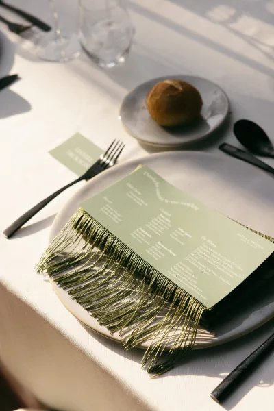 Wedding menu, a sage green menu card with tassel fringe, rests on a white tablecloth with soft sunlight shadows at the table.