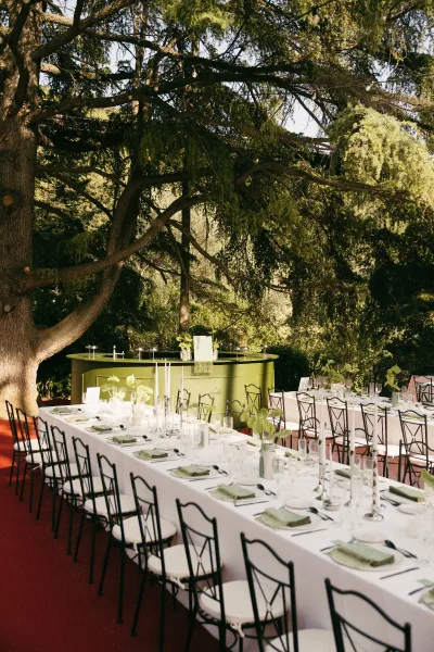 Reception tablescape for outdoor reception decor with taper candles set beneath garden trees and dense foliage over red flooring.