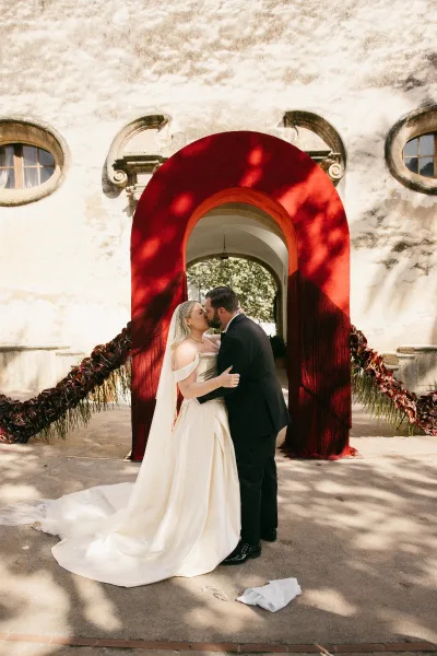 Wedding kiss portrait capturing a first kiss moment as the bride and groom embrace beneath a red ceremony arch by a sunlit stucco arched doorway.