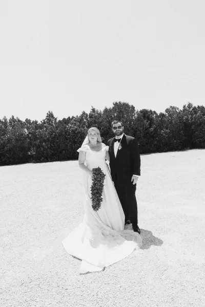 Couple portrait of a bride and groom standing, she holds a cascading bouquet as they face the camera in formal attire outdoors.