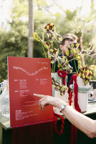 Wedding bar sign doubling as a signature cocktail sign, styled with anthuriums on an outdoor bar counter beneath sunlit garden trees