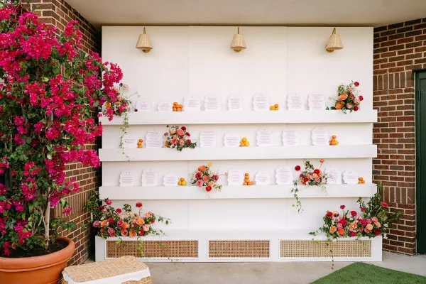 Wedding seating chart with escort card display on white shelving, accented by pink florals, greenery, and citrus against a brick wall backdrop