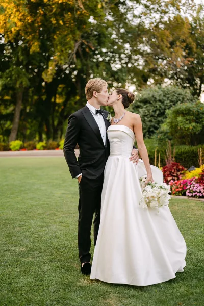 Wedding kiss portrait of bride and groom kissing on a garden lawn, her strapless dress and bouquet against his black tuxedo and bow tie