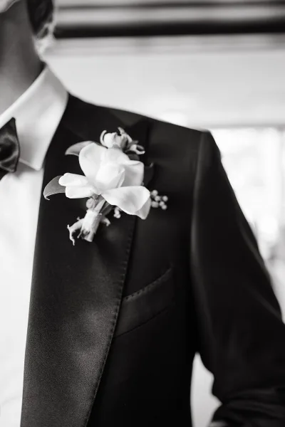 Groom boutonniere with a white orchid pinned to a black tuxedo satin lapel, bow tie and shirt visible against a blurred interior backdrop